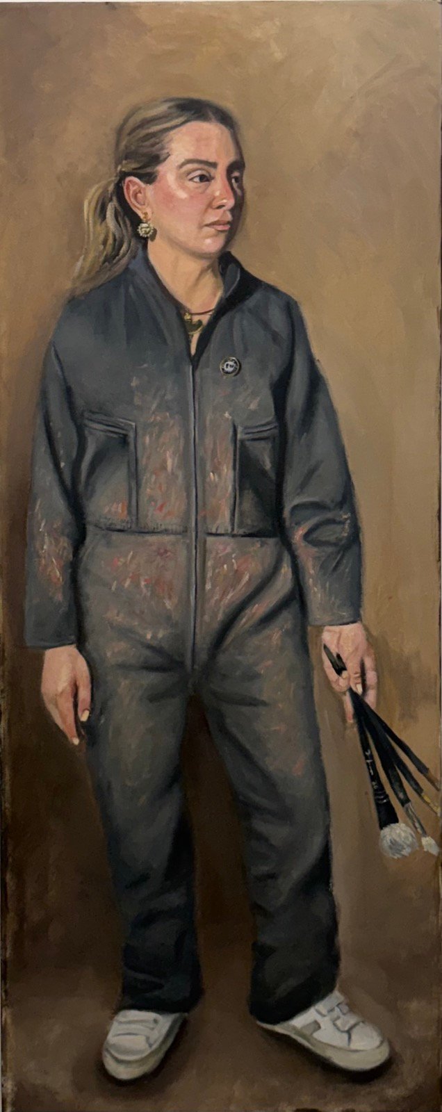 Full-length self-portrait of the artist standing in paint-stained grey coveralls, holding a fistful of brushes, against a warm brown ground